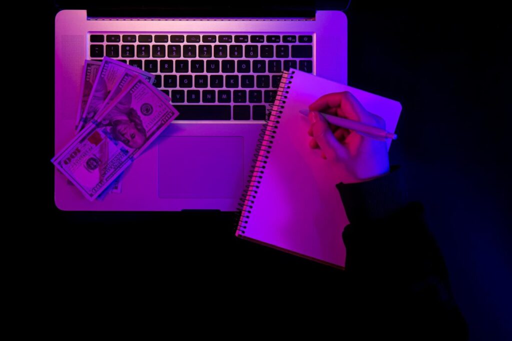 Laptop blank notepad and paper money in neon lighting top view